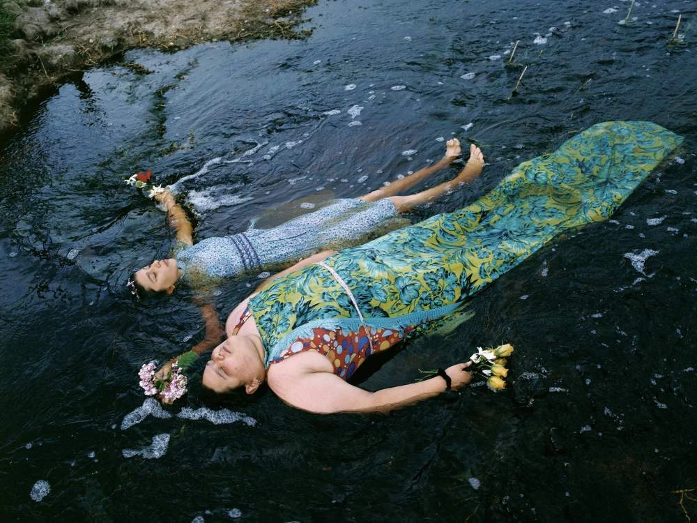 Due donne distese nell’acqua di un fiume con dei fiori tra le mani