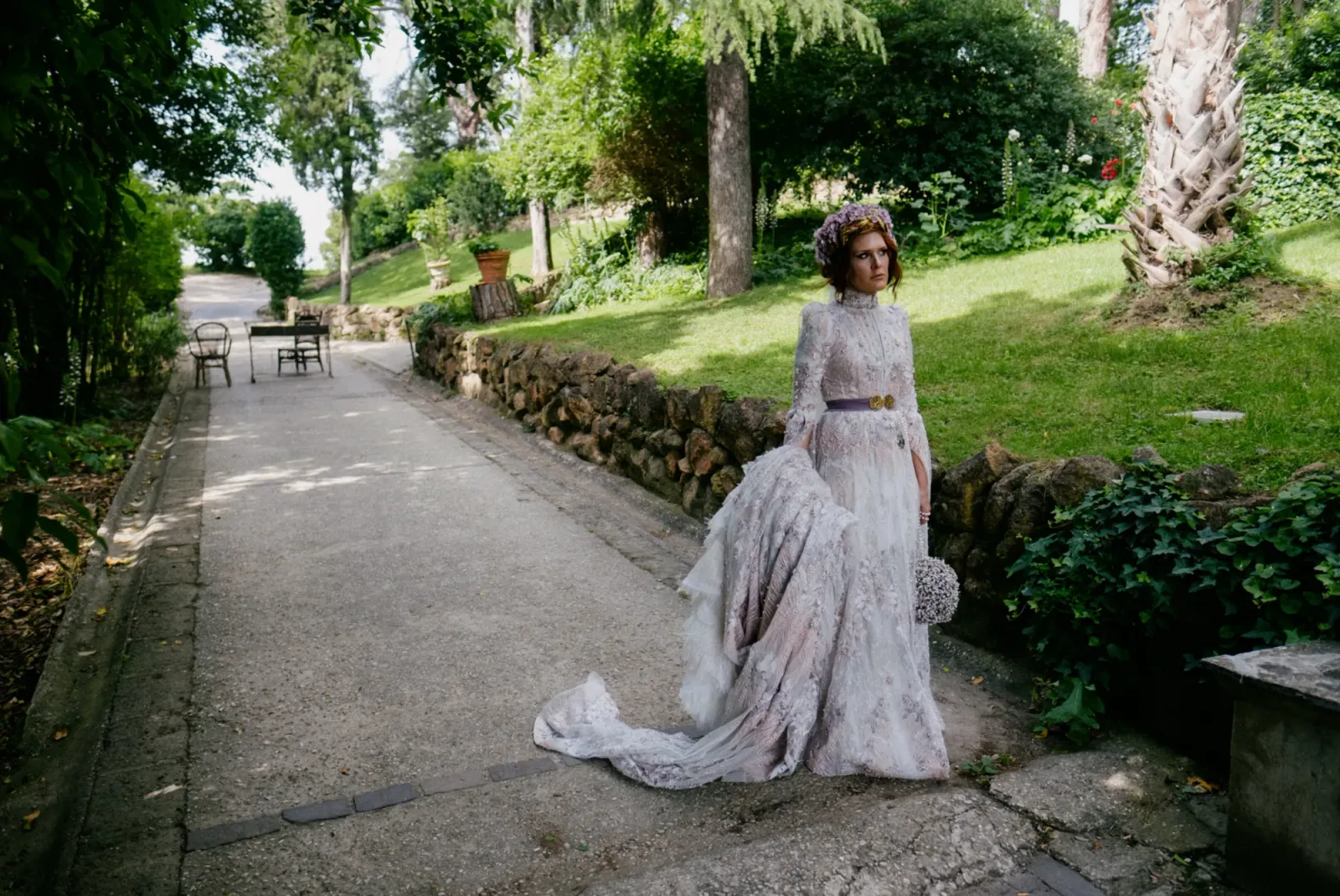 A bride in an embroidered lilac dress waiting on a garden path in full spring daylight