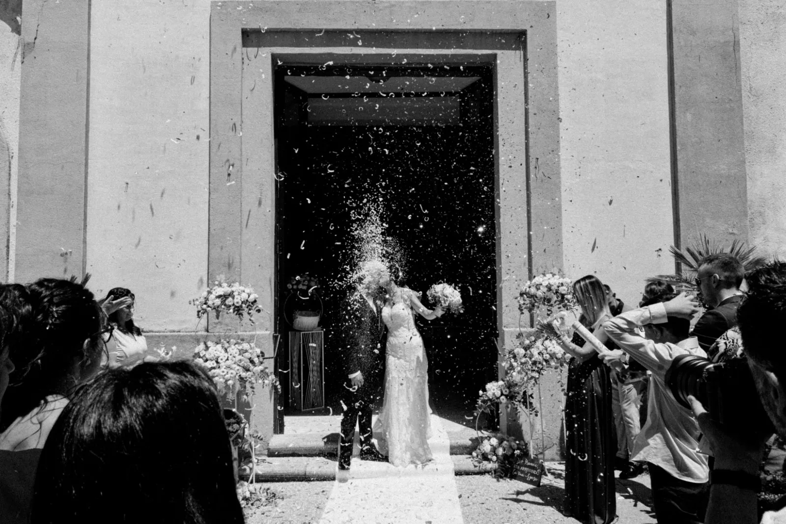 Confetti thrown over a couple in a church doorway under hard summer sun, in black and white