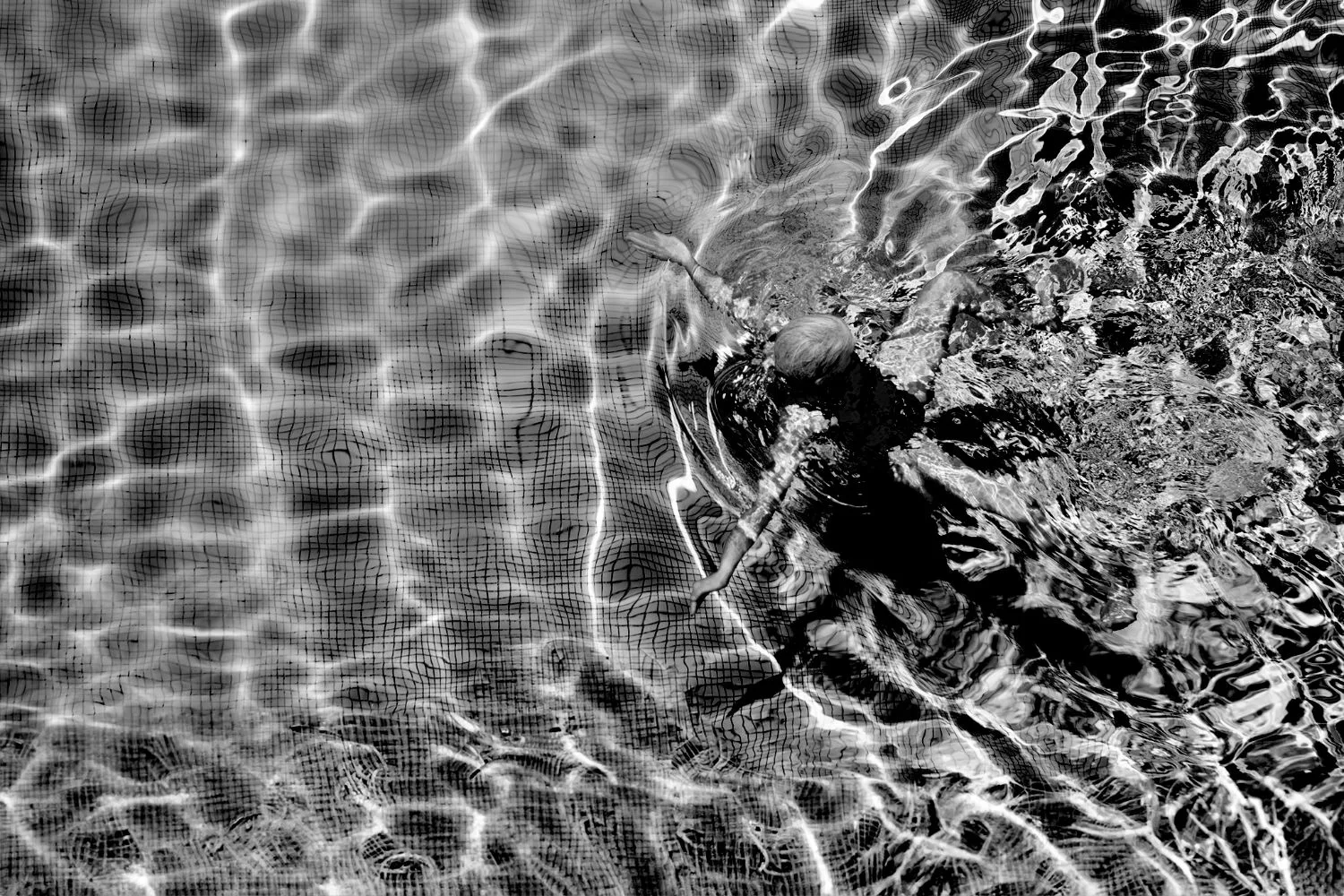 A swimmer seen through the rippling water of a tiled pool, in black and white