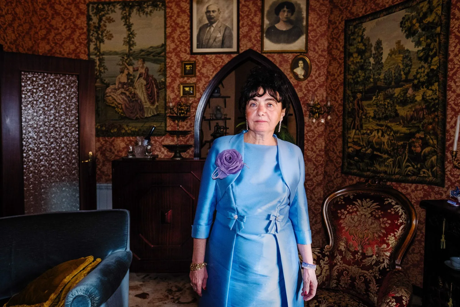 A guest in a blue dress standing in a front room hung with tapestries and family portraits