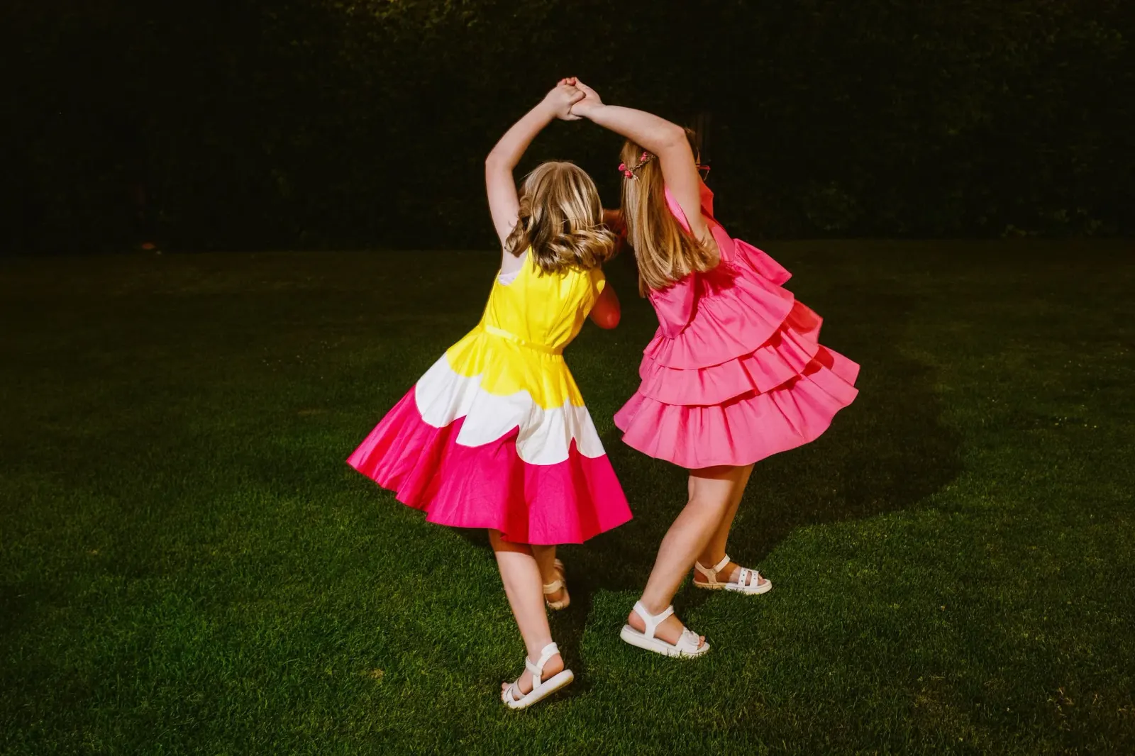 Two girls in yellow and pink dresses dancing on the grass at night