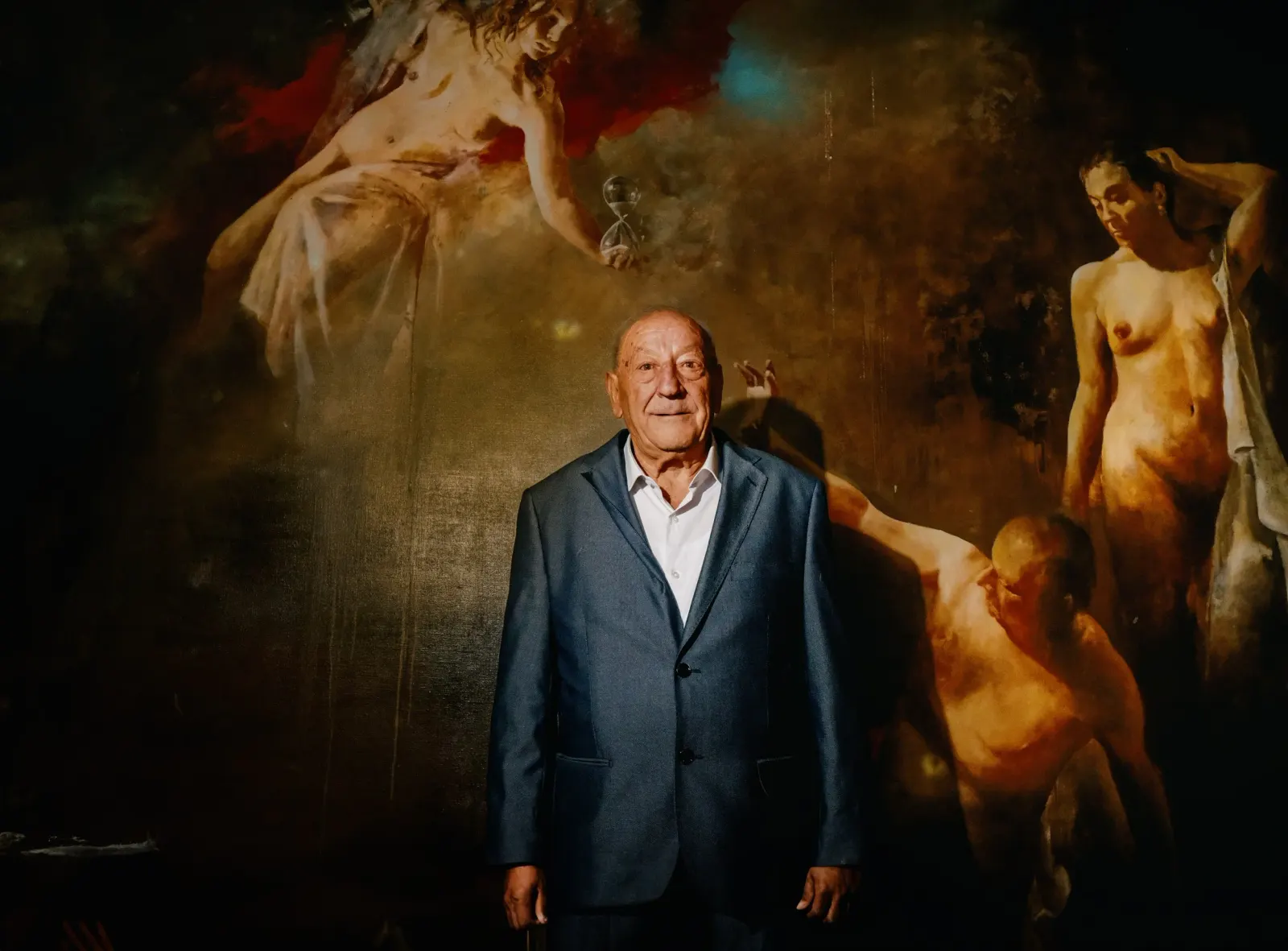 An elderly man in a suit standing in front of a painting of nudes