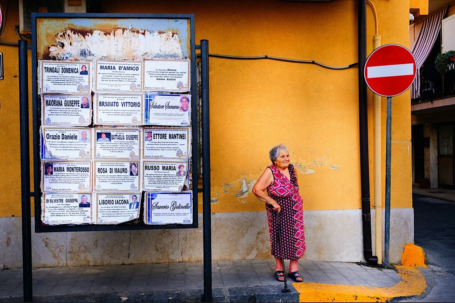 A woman standing beside a board of death notices on an orange wall