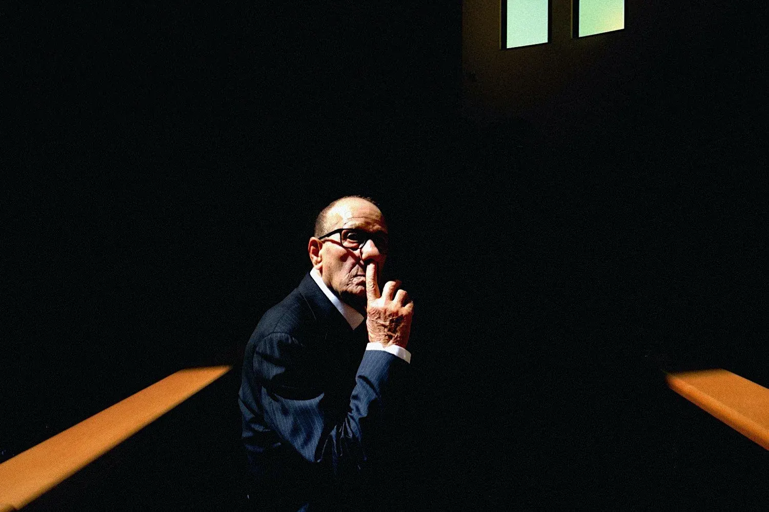 A man in glasses and a dark suit lit by a thin strip of light