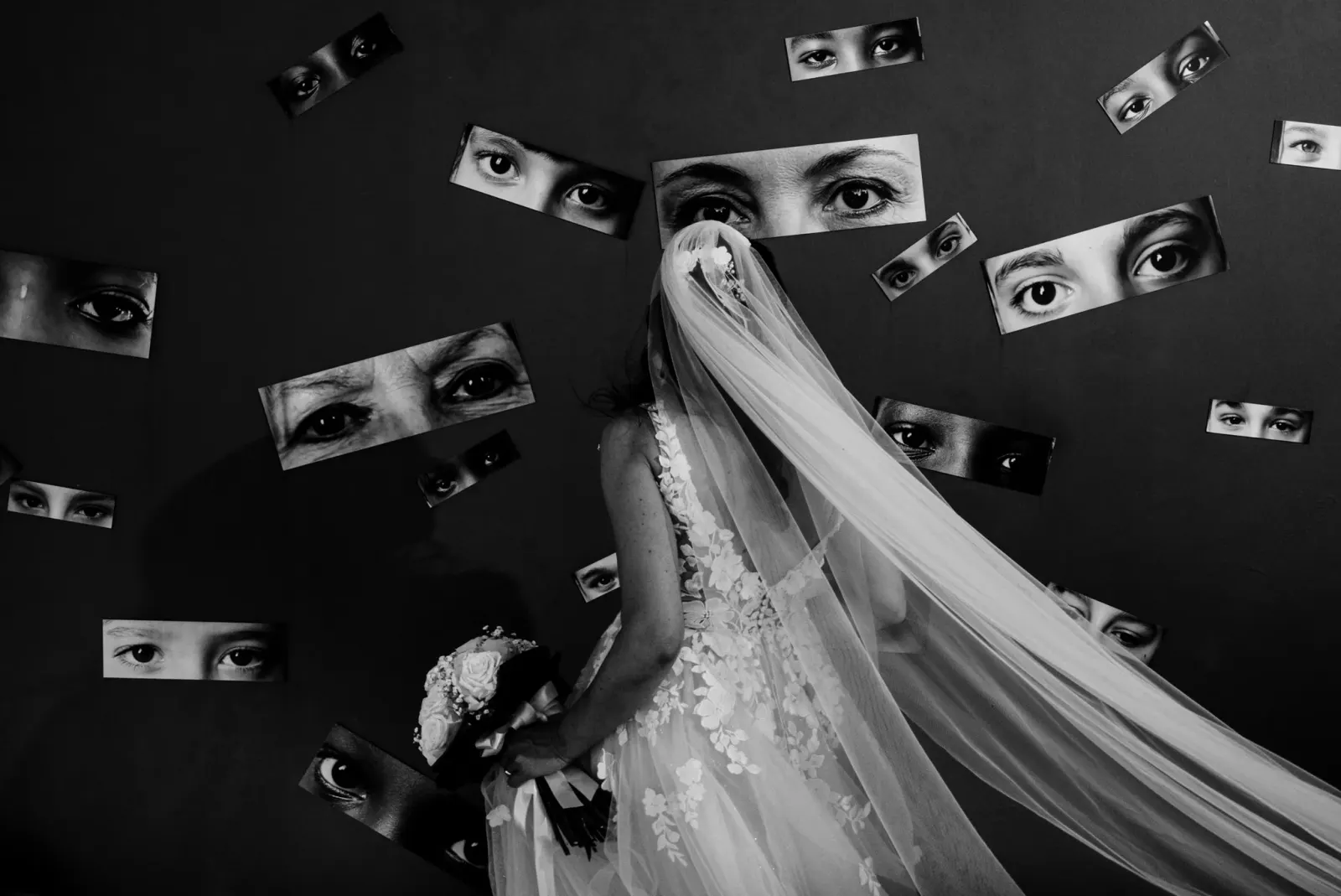 A bride lying beneath her veil, surrounded by cut out photographs of eyes