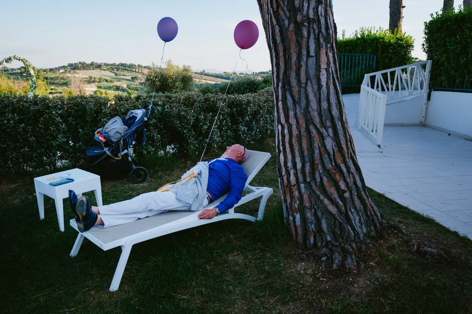 A man asleep on a sun lounger under a tree, two balloons tied nearby