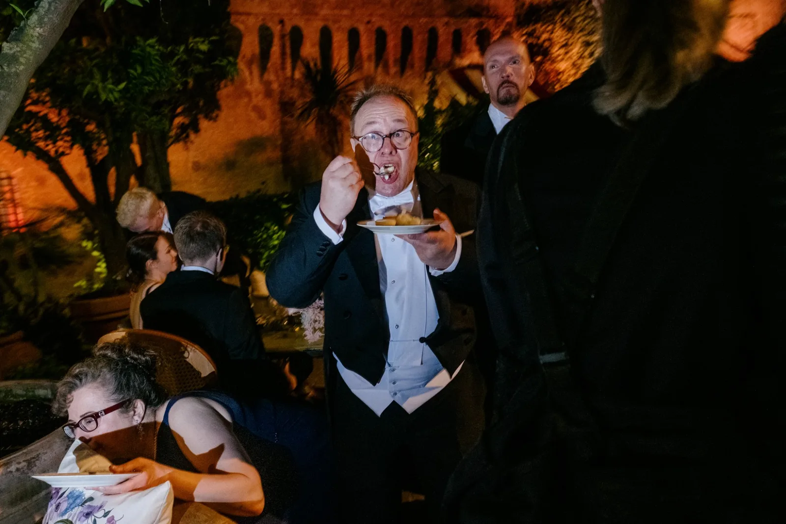 A guest in white tie eating from a plate at a night reception, another guest watching him over his shoulder