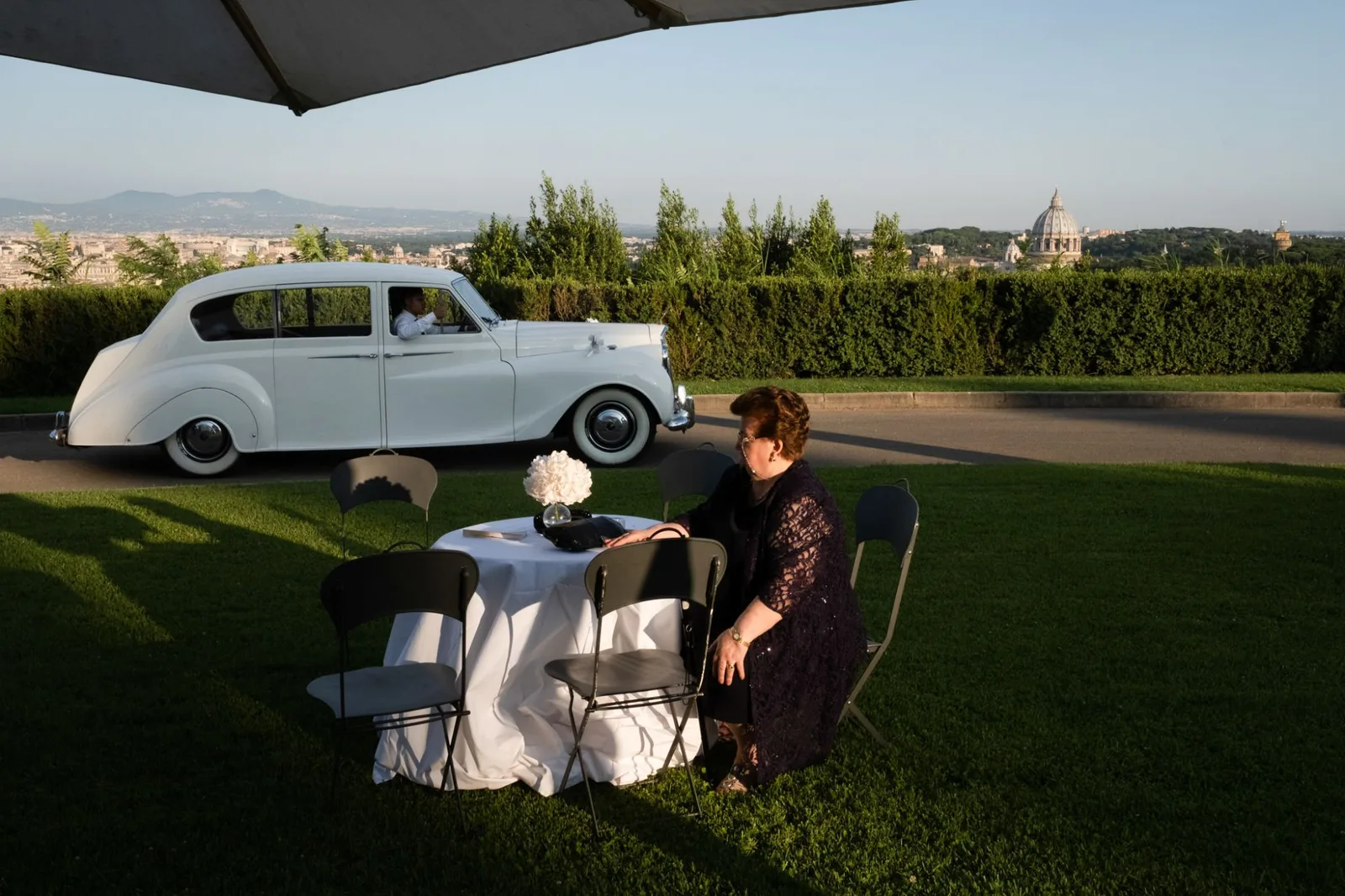 A vintage white car waiting on the drive while a guest sits alone at a table, the dome of St Peter’s on the horizon