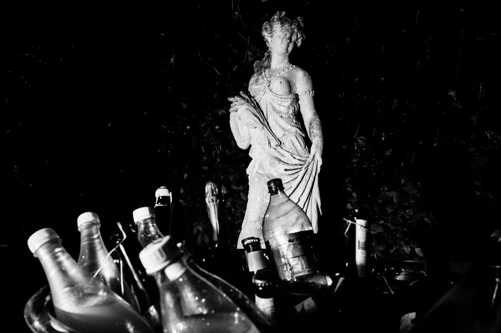 Bottles crowded on a tray in front of a garden statue after dark, in black and white