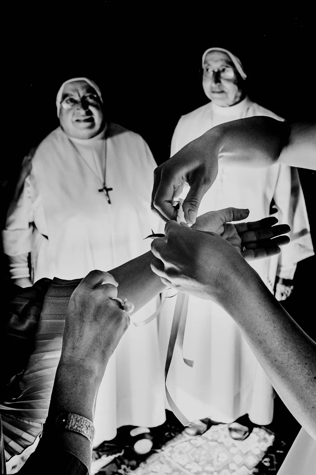 Two nuns watching a ribbon being tied around a wrist, in black and white