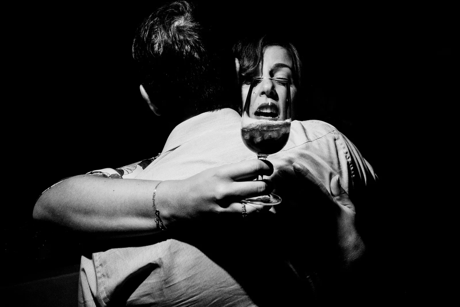 Two guests holding on to each other, one face seen through the wine glass in her hand, in black and white