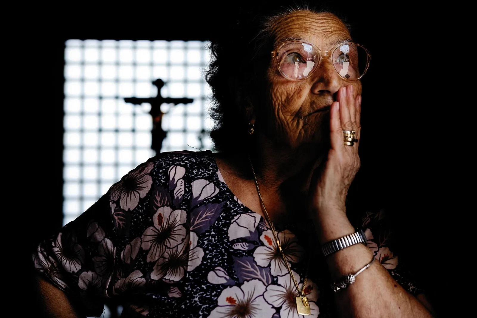 An elderly woman with a hand to her face in a Sicilian church, a crucifix behind her