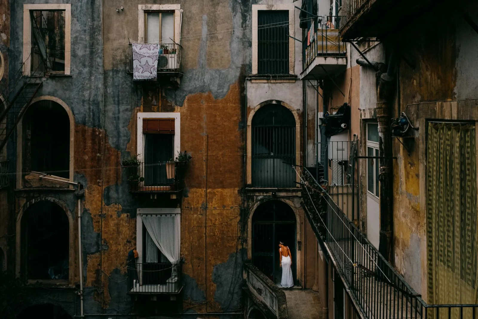 A bride alone on a landing in a Catania courtyard of peeling balconies and washing lines