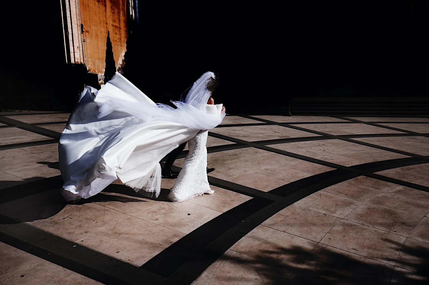 A bride and groom crossing a sunlit square, her dress and veil thrown sideways by the wind