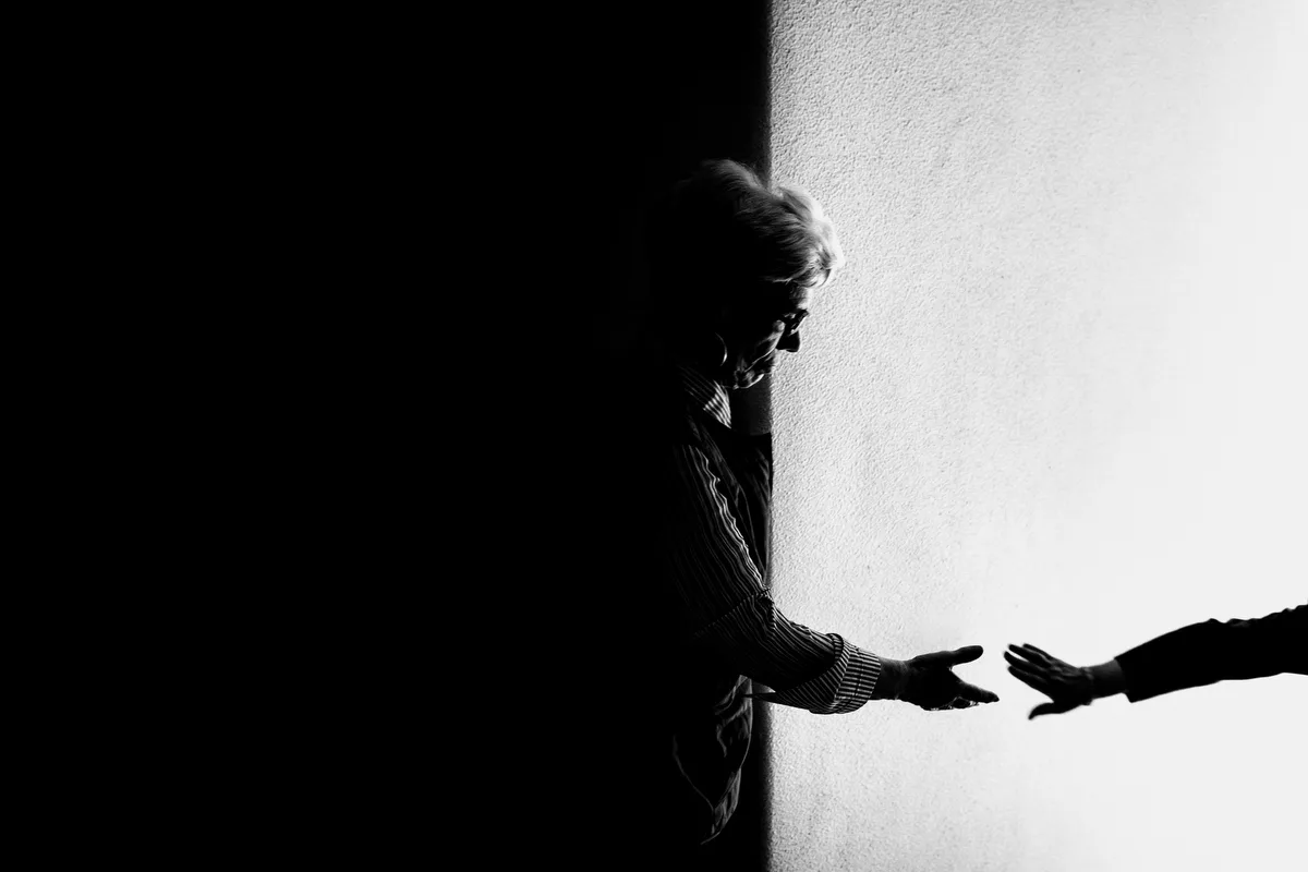 An elderly woman in shadow reaching her hand towards another hand across a white wall, in black and white